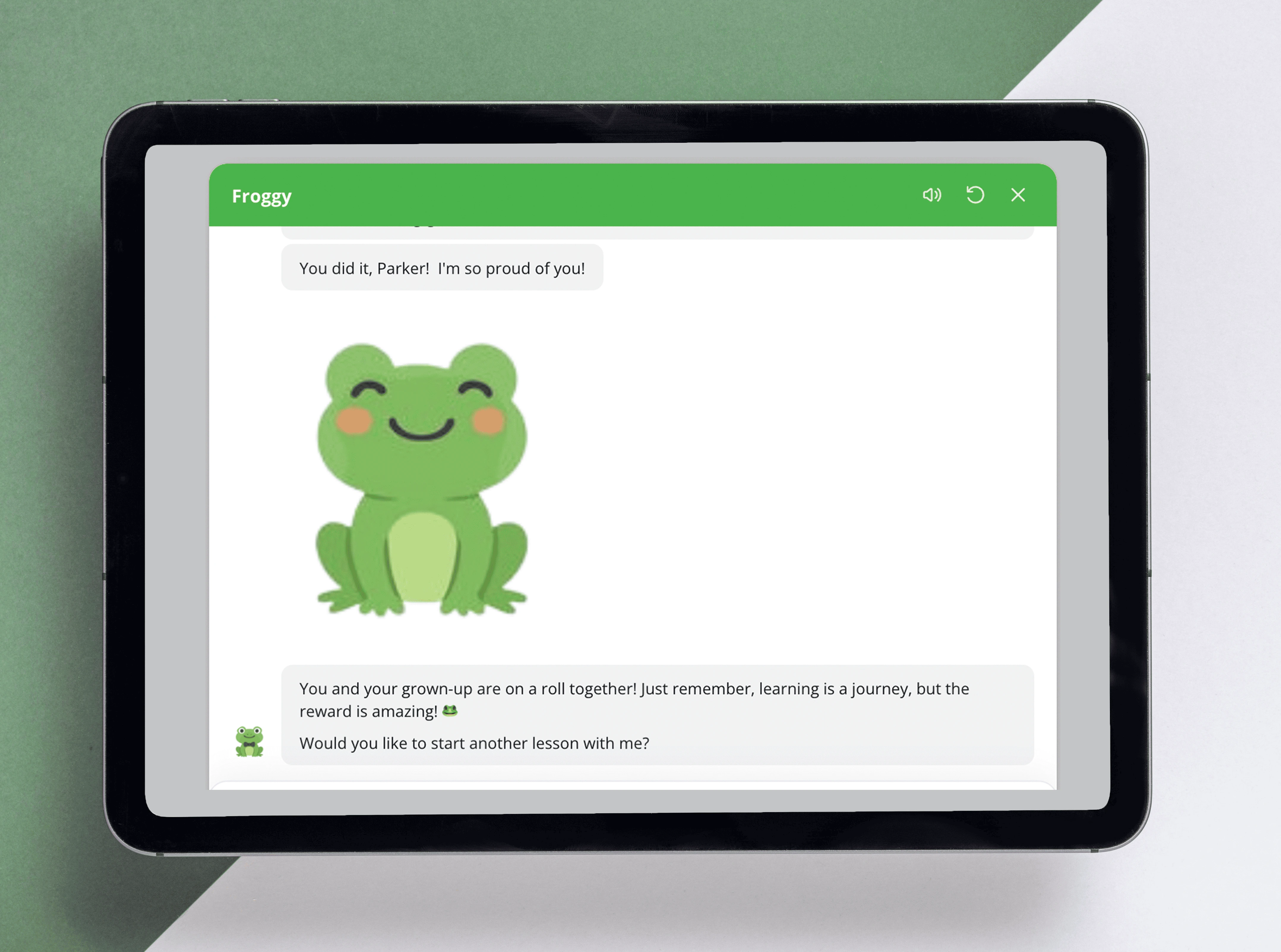 Tablet displaying a children's learning app with a smiling cartoon frog character and a congratulatory message celebrating lesson completion, along with a prompt to start another lesson.