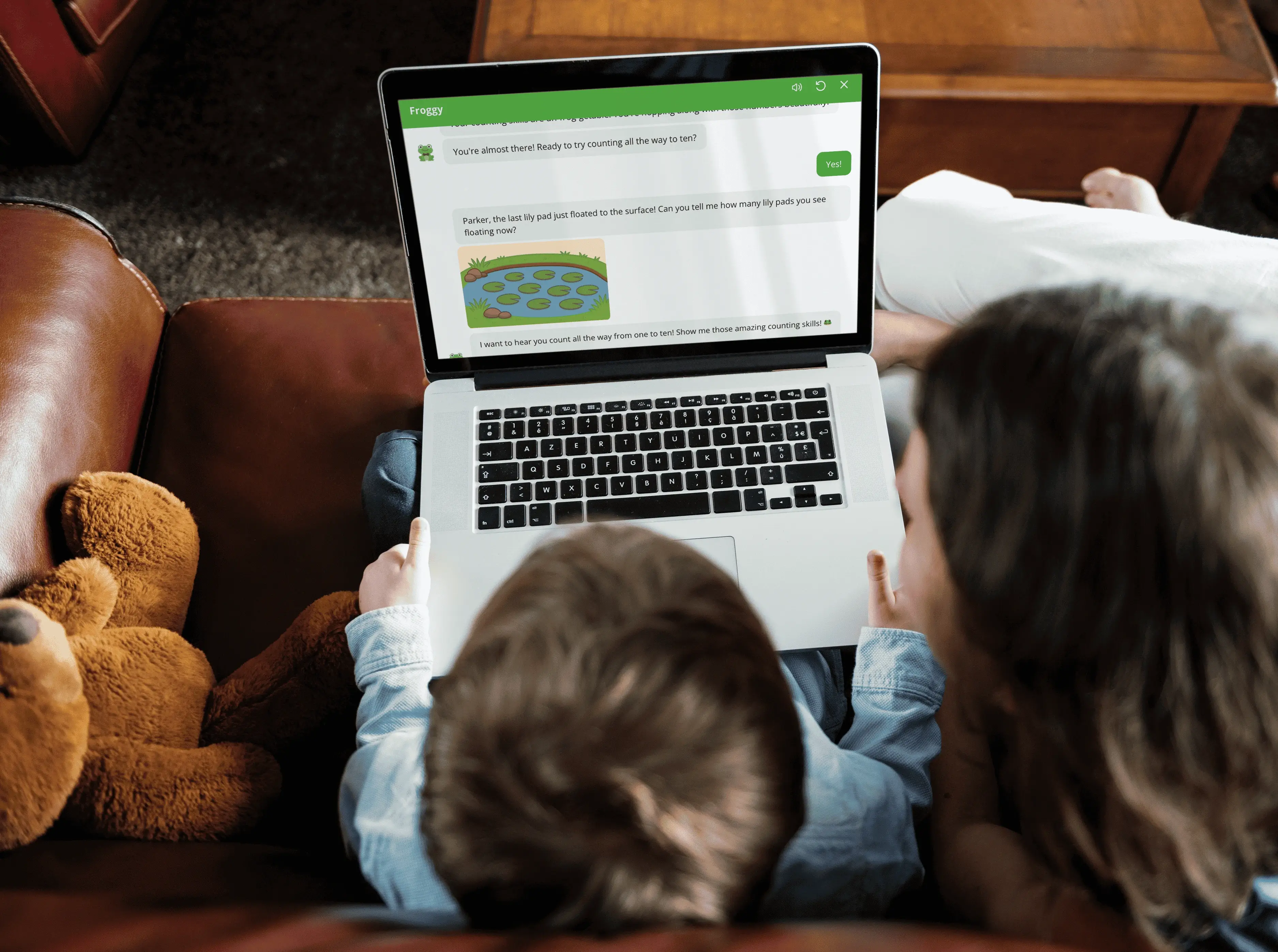 Overhead view of a child and an adult sitting
                                on a couch using a laptop, with a learning app on
                                screen showing a counting activity with lily pads,
                                and a teddy bear nearby.