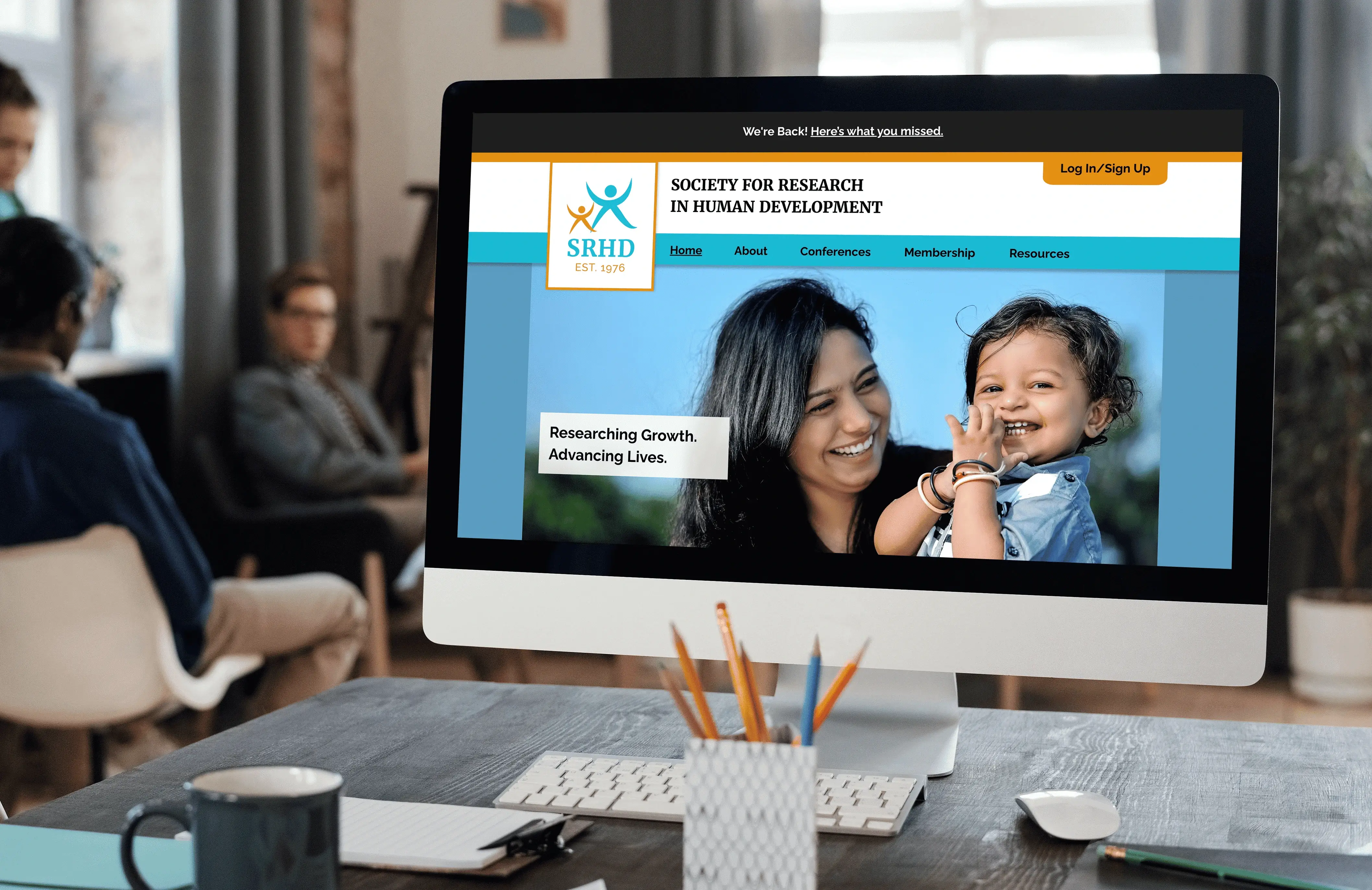 A desktop display featuring the SRHD website,
                                the desk holding the display is in front of a group
                                of slightly blurred people sitting in the
                                background.